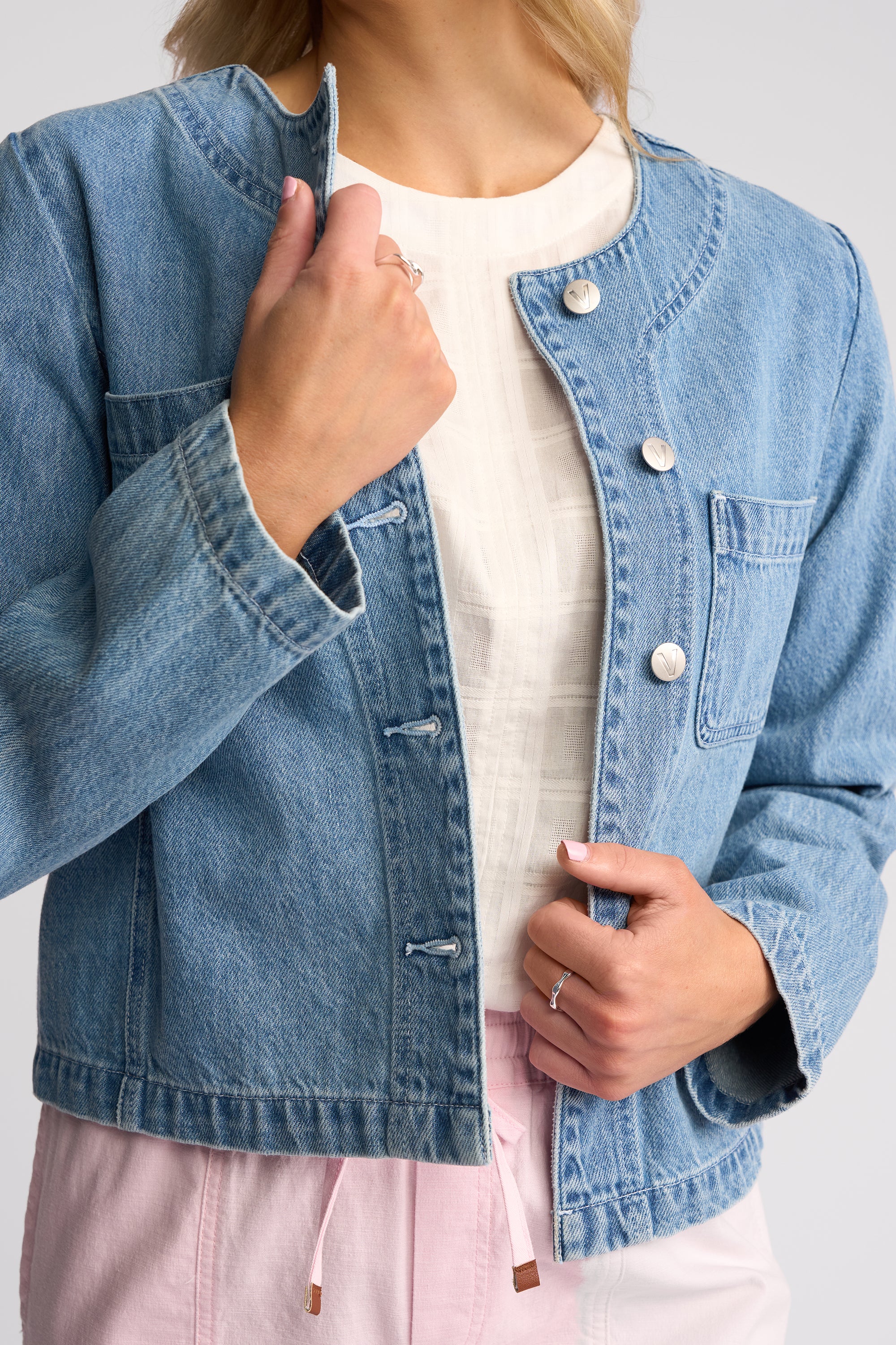 A woman wearing a cropped light blue denim jacket over a white top and pink drawstring pants, shown from the shoulders to the hips against a light gray studio background.