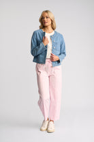 A woman modeling a light blue cropped denim jacket, pink drawstring pants, a white top, and beige sneakers against a light gray studio background.