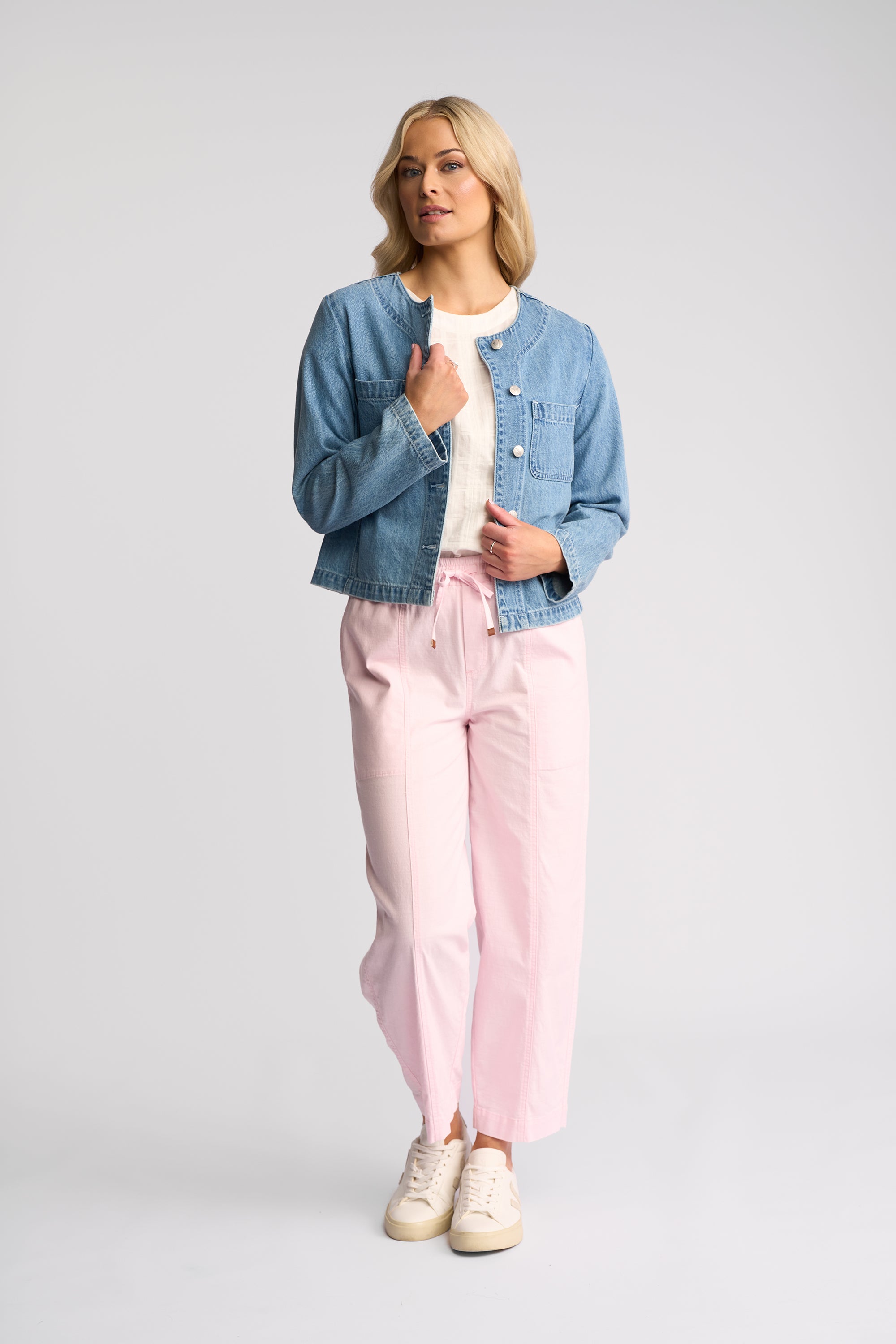 A woman modeling a light blue cropped denim jacket, pink drawstring pants, a white top, and beige sneakers against a light gray studio background.