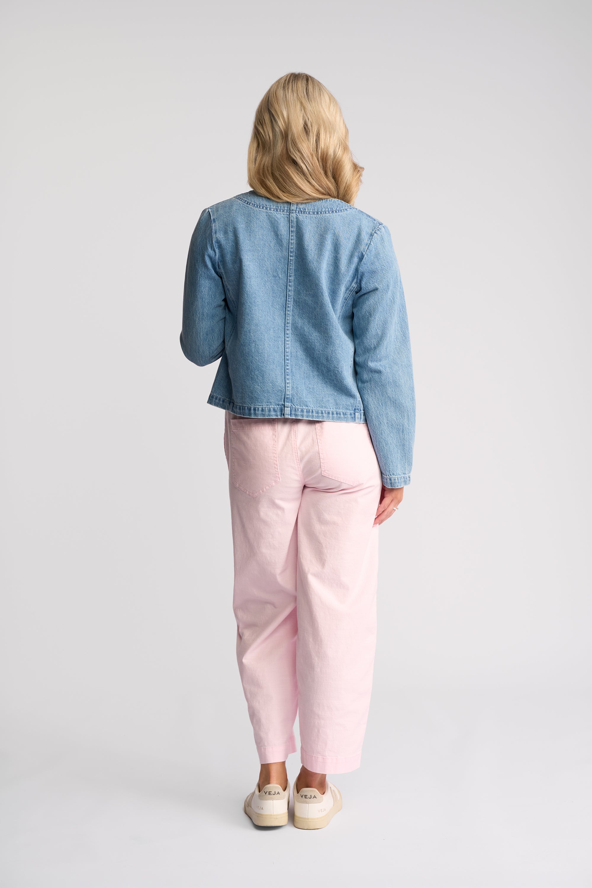 A woman viewed from behind wearing a cropped light blue denim jacket, light pink trousers, and white sneakers against a plain light gray studio background.