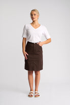 A woman modeling a white short-sleeve V-neck t-shirt and a dark brown knee-length skirt, paired with white slide sandals, against a light gray studio background.