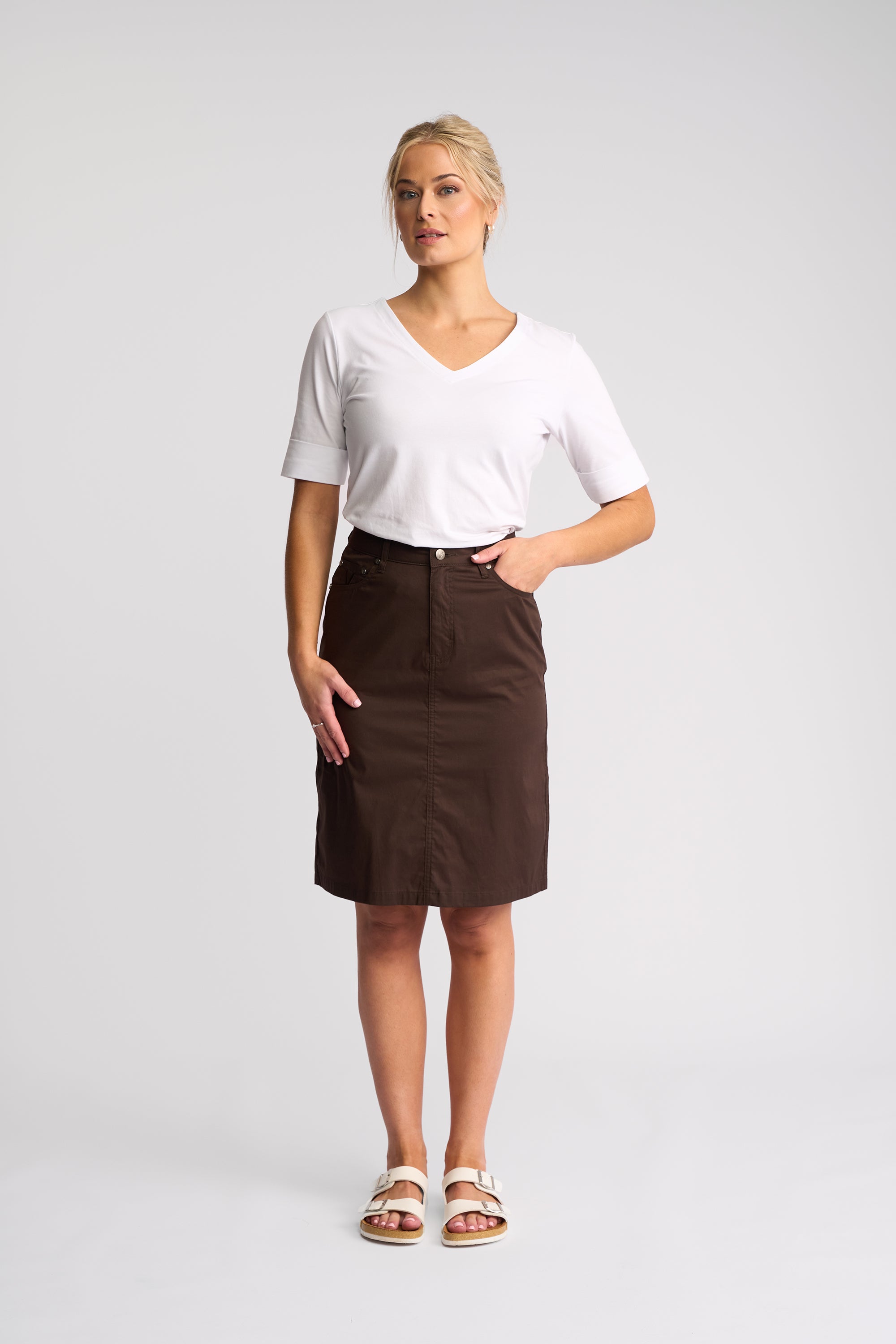 A woman modeling a white short-sleeve V-neck t-shirt and a dark brown knee-length skirt, paired with white slide sandals, against a light gray studio background.
