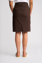 A woman shown from the back wearing a dark brown knee-length skirt with a front button closure and side pockets, paired with a white top and beige sandals, against a light gray studio background.