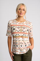 A woman modeling a short-sleeve crewneck top with a multicolor botanical and tribal pattern on a cream background, paired with olive green pants, against a light gray studio background.
