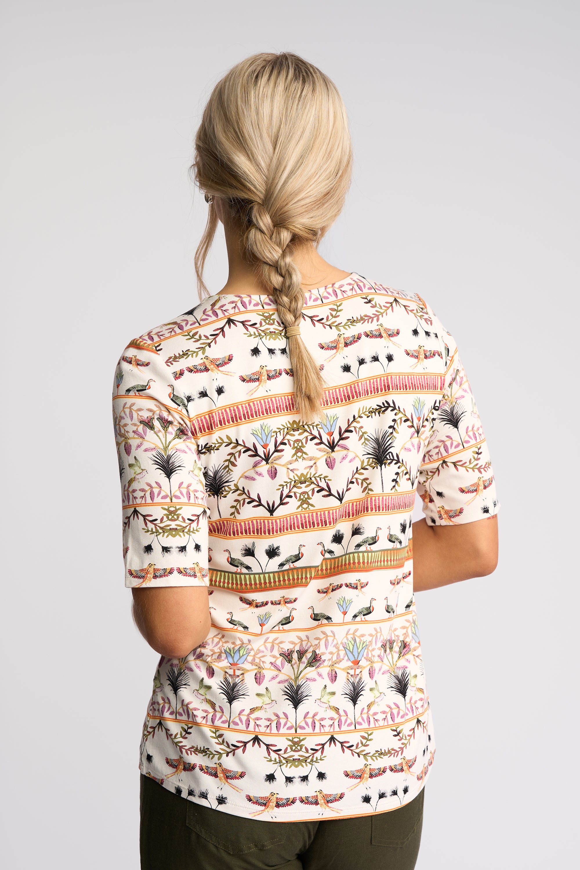 A woman viewed from behind wearing a short-sleeve top with a colorful botanical and geometric pattern on a cream background, paired with dark olive pants, against a light gray studio background.