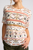 A woman wearing a short-sleeve crewneck top with a colorful botanical and bird pattern on a white base, paired with dark olive pants, against a light gray studio background.
