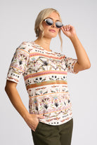 A woman wearing a short-sleeve crewneck top with a colorful horizontal patterned design, paired with dark olive pants and sunglasses, against a light gray studio background.