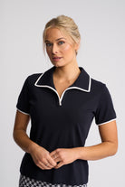 A woman modeling a short-sleeve polo shirt in navy blue with white piping trim on the collar and sleeve cuffs, set against a plain light gray studio background.