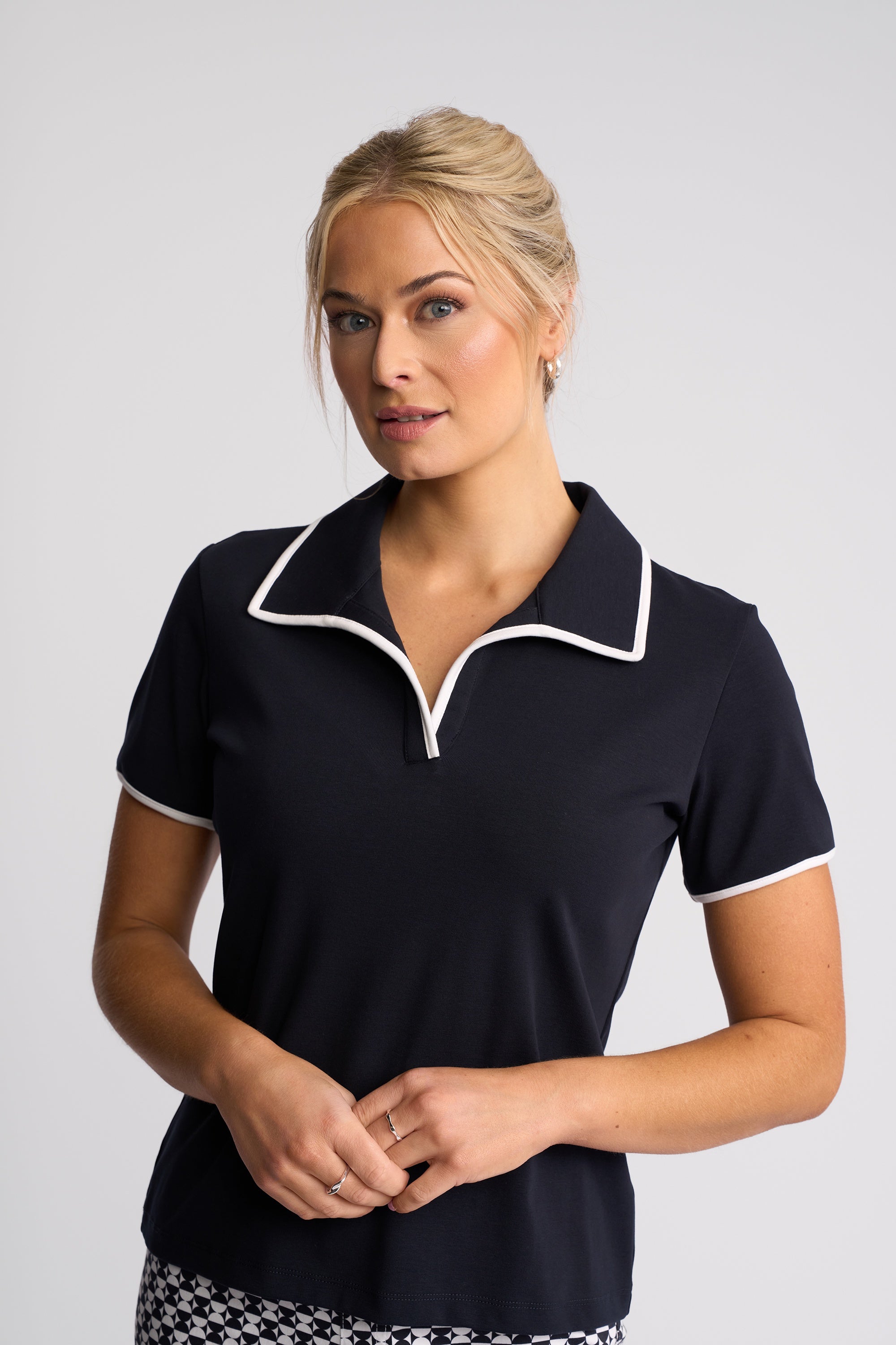 A woman modeling a short-sleeve polo shirt in navy blue with white piping trim on the collar and sleeve cuffs, set against a plain light gray studio background.