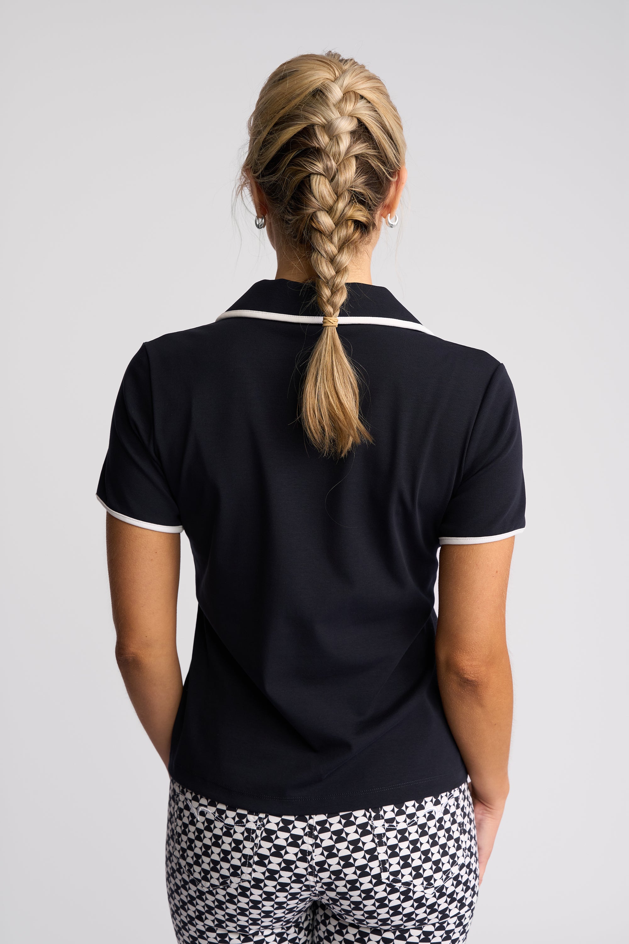 A woman viewed from behind wearing a navy blue short-sleeve polo shirt with white trim on the collar and sleeve cuffs, paired with black and white patterned pants, against a light gray studio background.