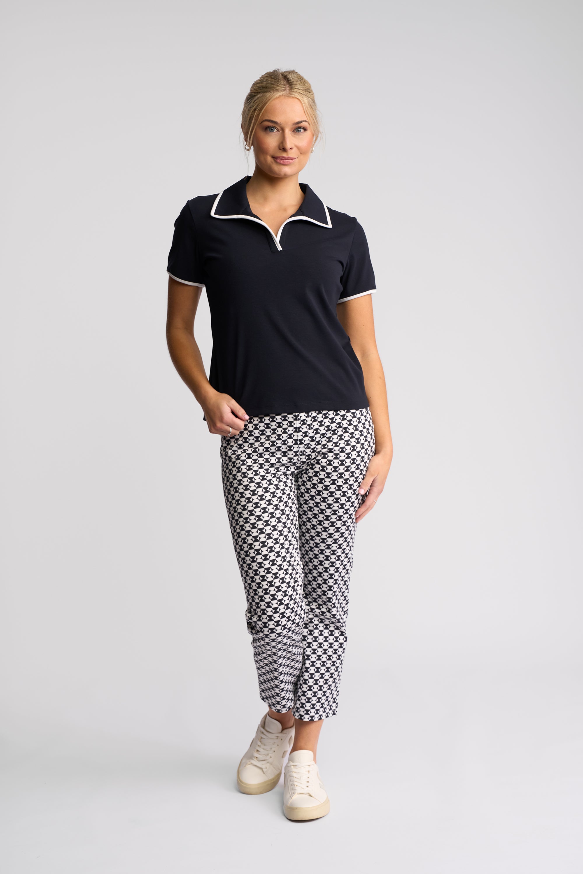 A woman modeling a short-sleeve polo shirt and patterned trousers against a light gray studio background. The polo is black with white piping, and the trousers have a black-and-white houndstooth pattern.