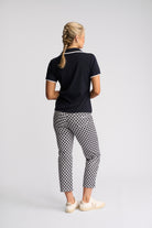 A woman is shown from behind wearing a black short-sleeve polo shirt with white trim, black and white checkered cropped trousers, and white sneakers, set against a plain light gray studio background.
