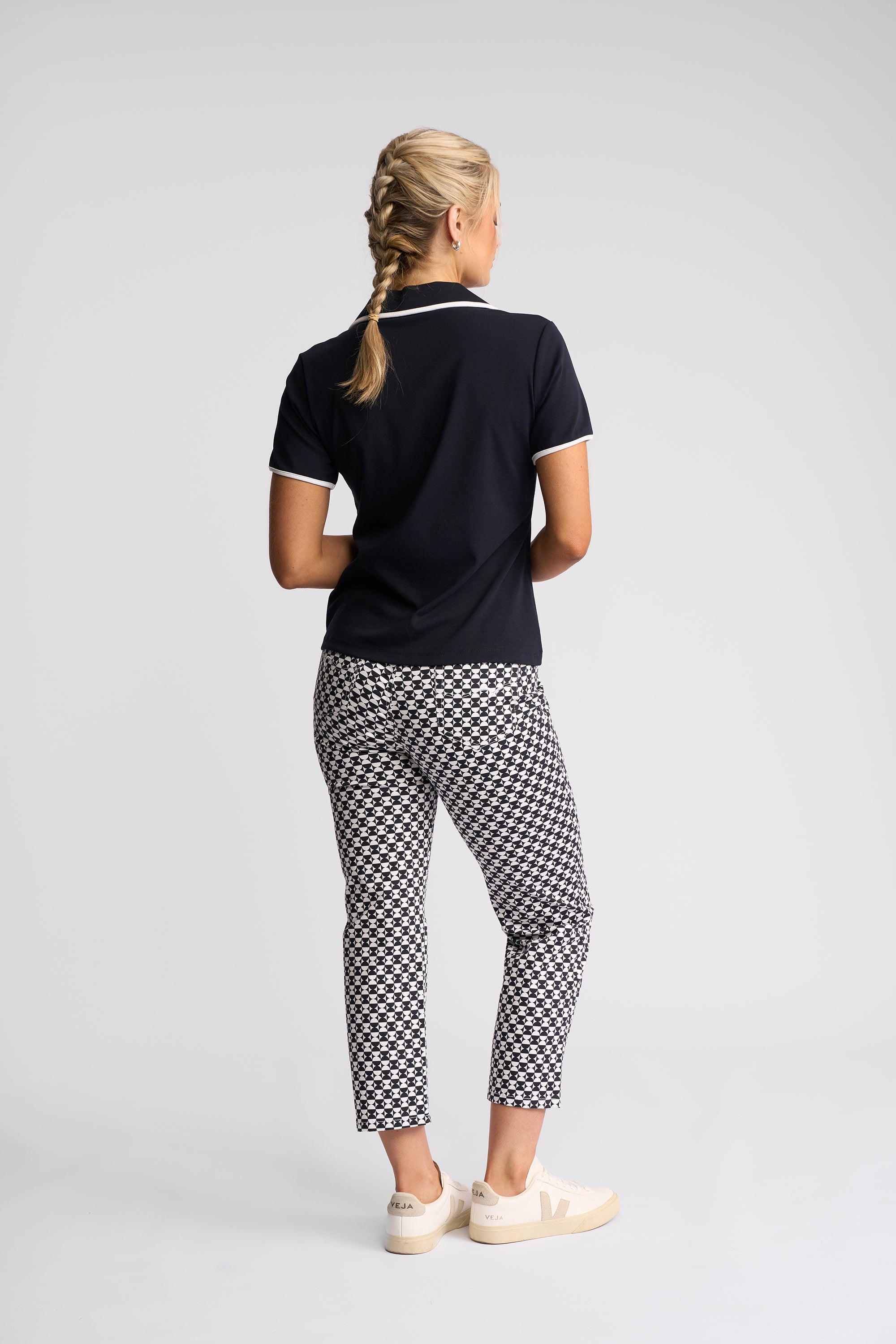 A woman is shown from behind wearing a black short-sleeve polo shirt with white trim, black and white checkered cropped trousers, and white sneakers, set against a plain light gray studio background.