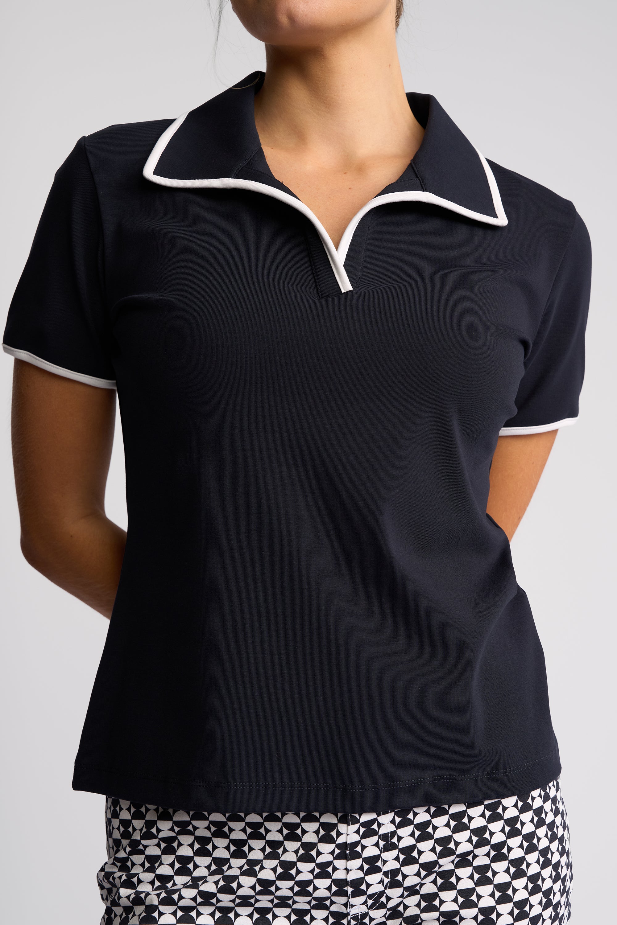 A woman wearing a short-sleeve polo shirt in black with white piping trim on the collar and sleeve cuffs, paired with a black and white patterned skirt, against a light gray studio background.