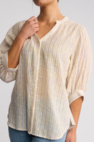 A woman wearing a cream-colored blouse with thin vertical blue stripes and small white embroidered details, paired with blue jeans against a light gray studio background.