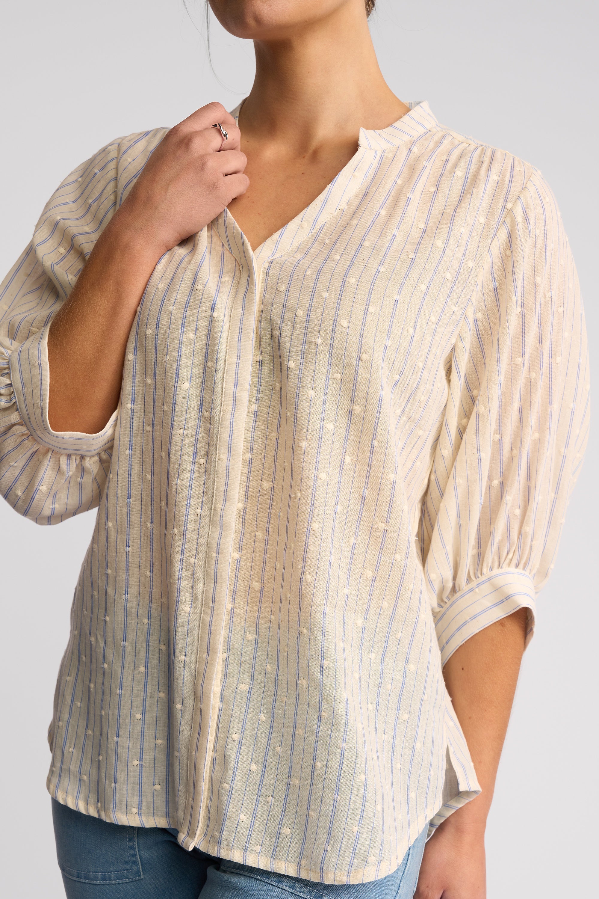 A woman wearing a cream-colored blouse with thin vertical blue stripes and small white embroidered details, paired with blue jeans against a light gray studio background.