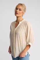 A woman wearing a light beige and white vertically striped button-up blouse with puff sleeves, paired with blue jeans, photographed against a solid light gray background.