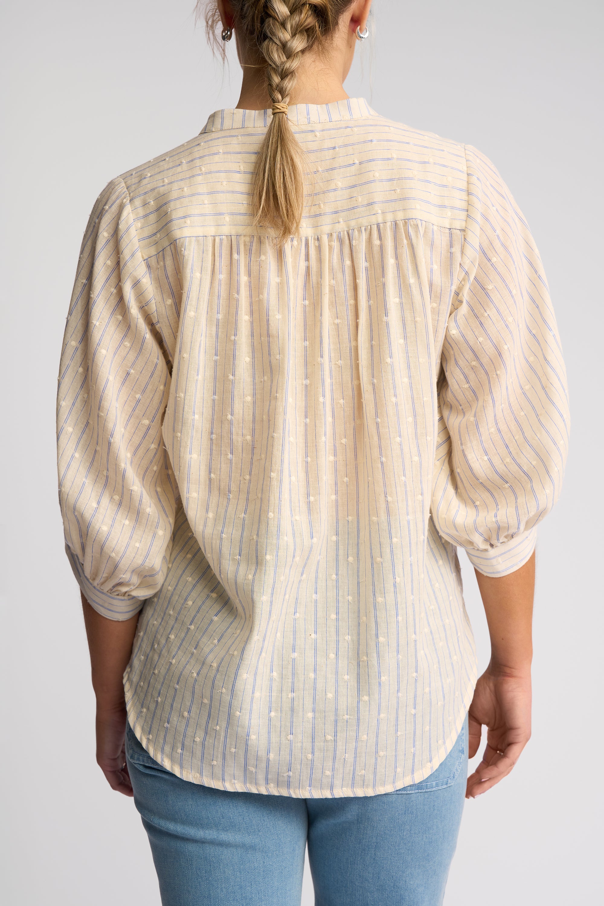 A woman viewed from the back wearing a cream-colored blouse with thin vertical stripes and light blue jeans, set against a plain light gray background.