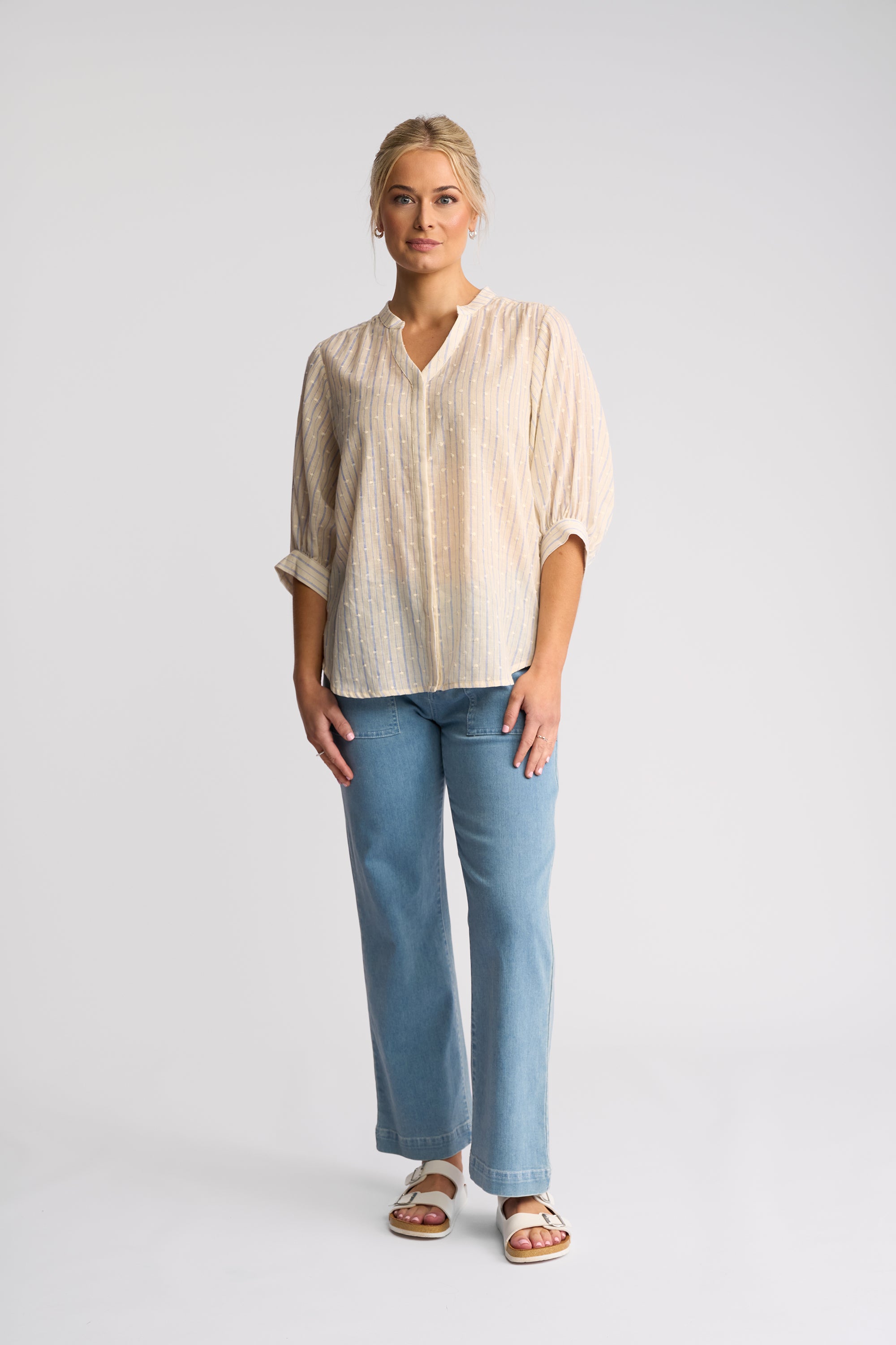A woman modeling a light beige and white vertically striped short-sleeve blouse with rolled cuffs, paired with light blue wide-leg jeans and beige sandals, against a light gray studio background.