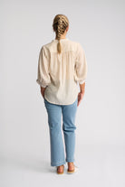 A woman viewed from behind wearing a cream and light blue vertically striped blouse with puff sleeves, paired with light blue jeans and white slide sandals, against a light gray studio background.