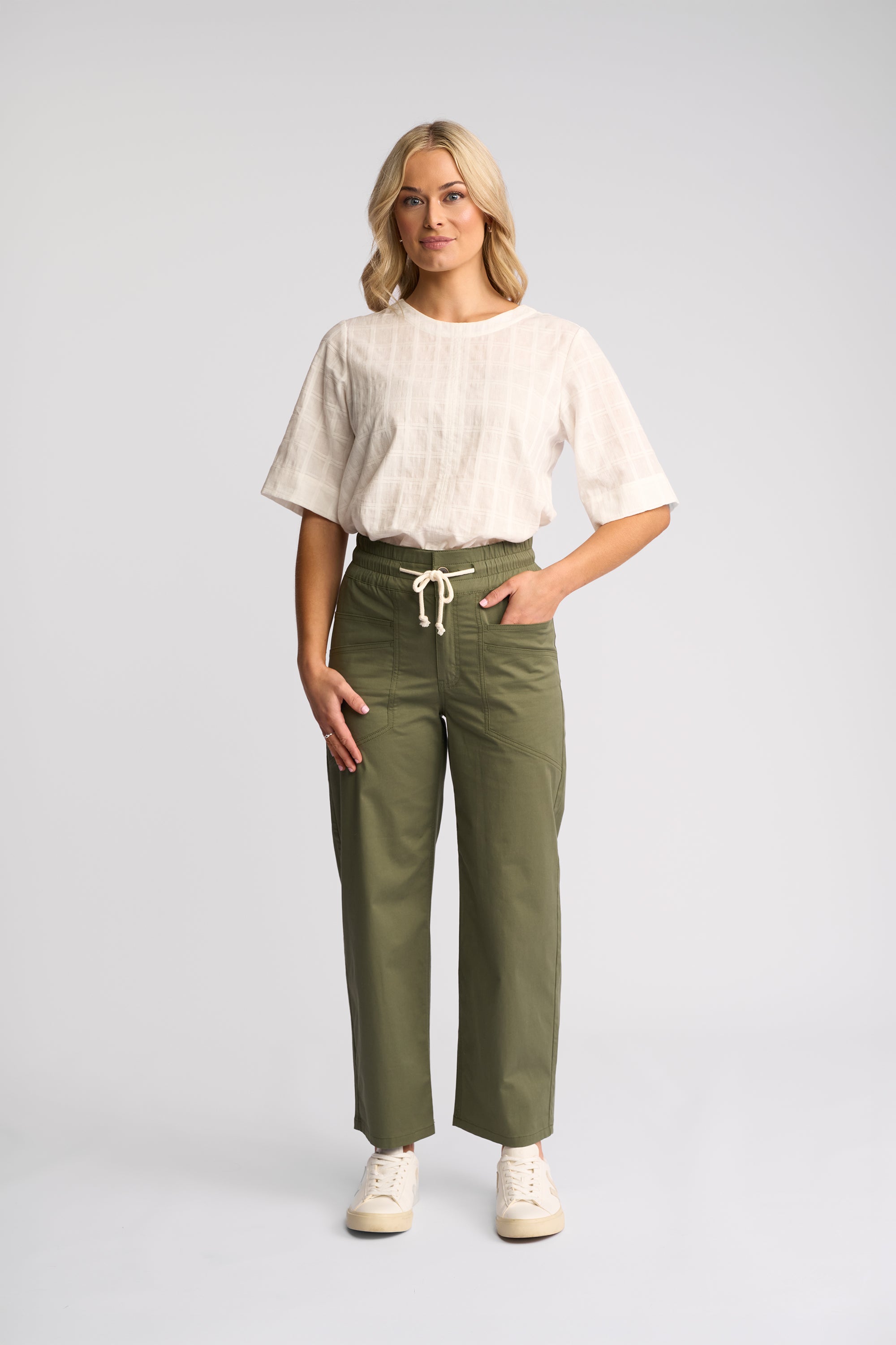 A woman modeling a pair of olive green wide-leg trousers with a white drawstring, paired with a white short-sleeve top and white sneakers, against a light gray studio background.