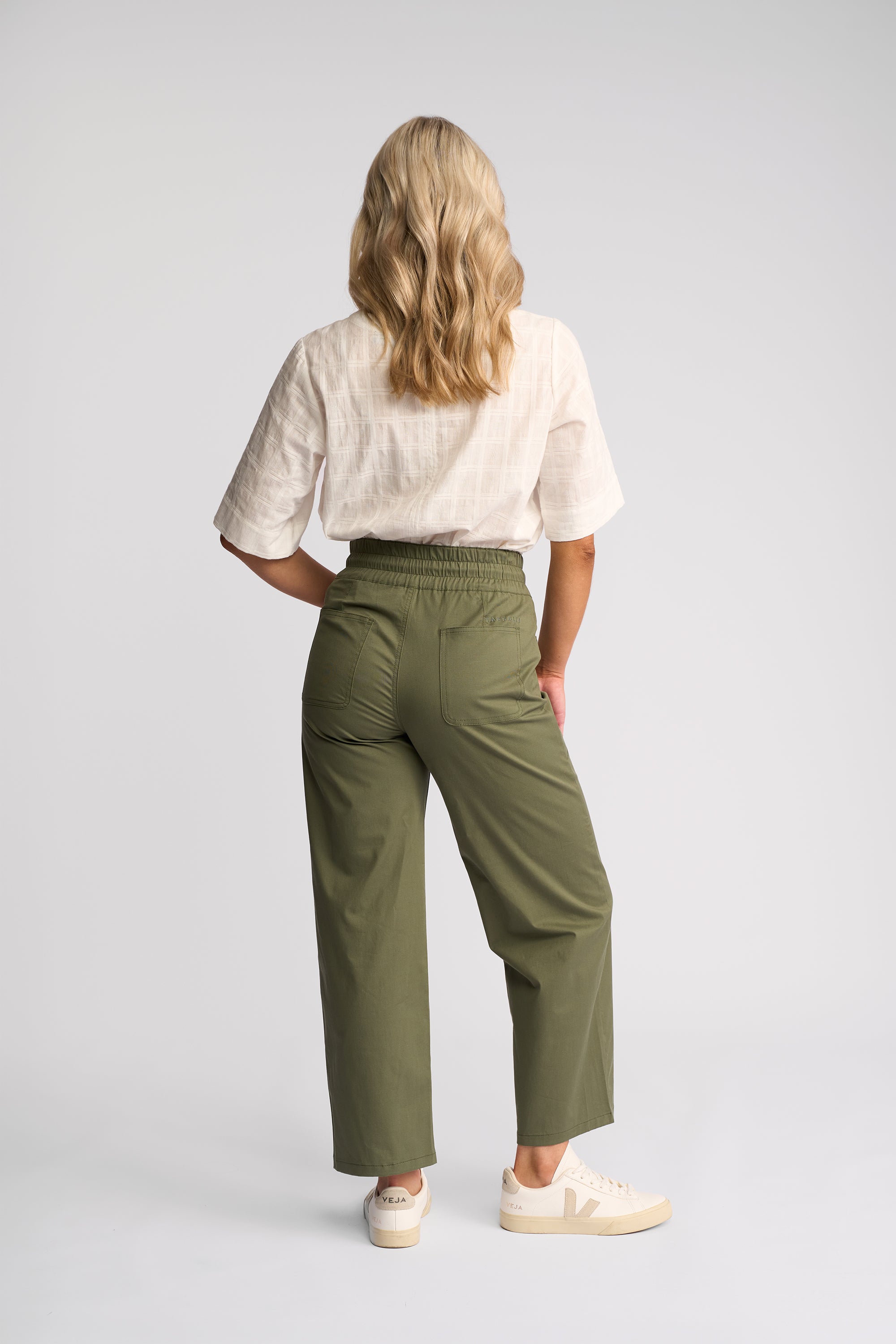 A woman shown from the back wearing olive green wide-leg cropped pants, a white short-sleeve top, and white sneakers against a light gray studio background.