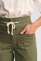 Close-up of a person wearing olive green drawstring pants with a side pocket and a cream-colored top tucked in, against a light gray background.