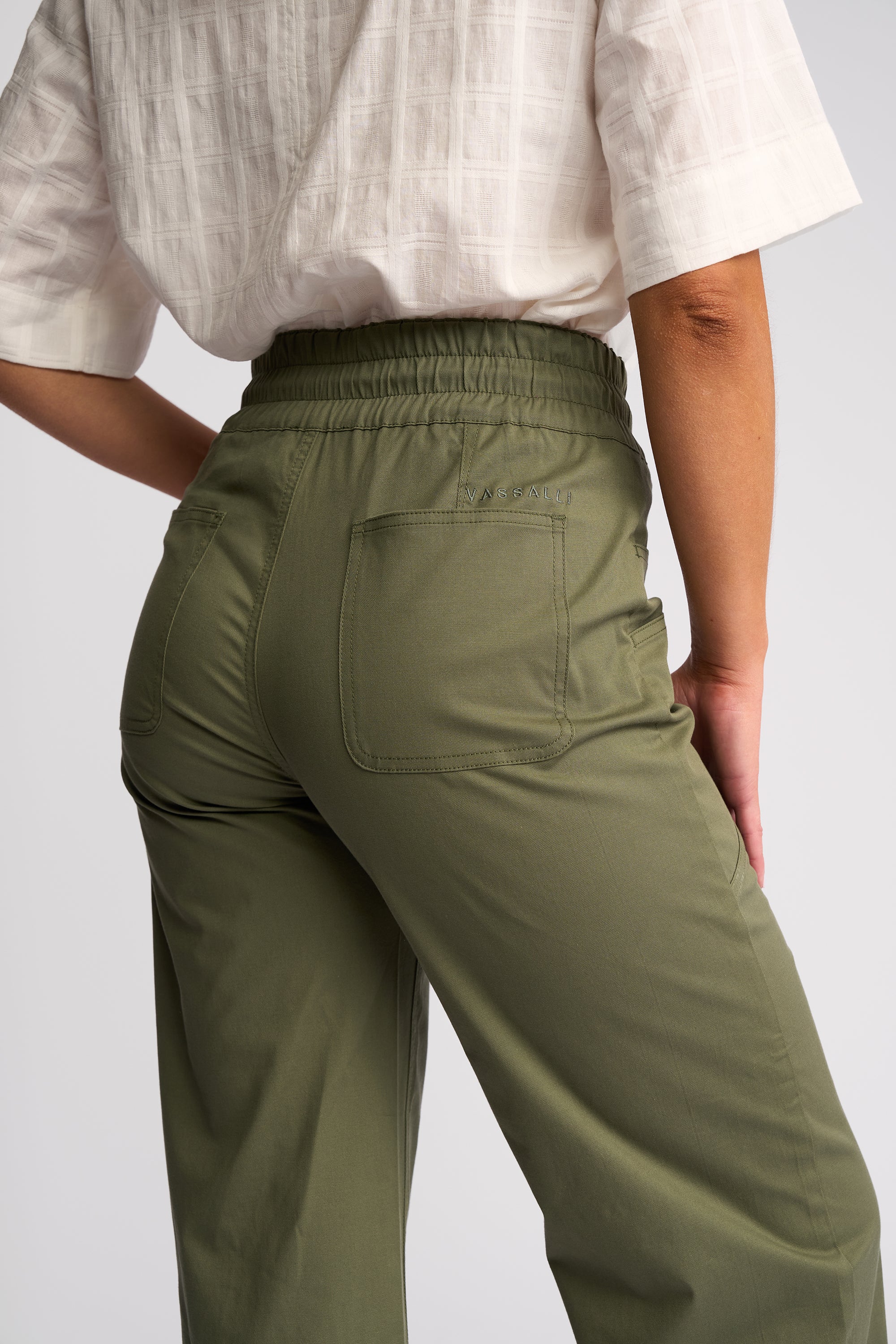 A close-up back view of a person wearing olive green wide-leg trousers with an elastic waistband and a white short-sleeve top, against a light gray studio background.