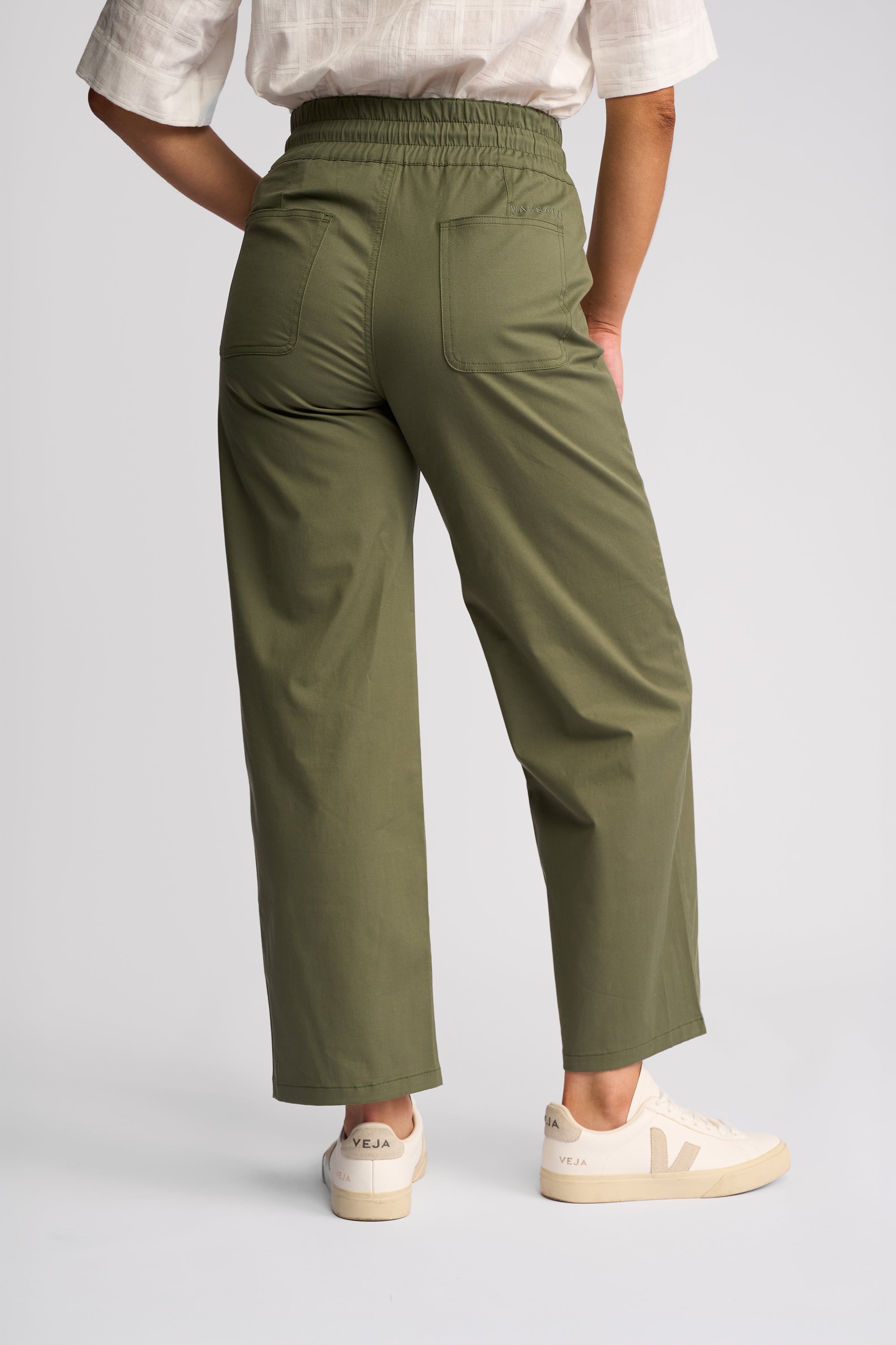 A person shown from the back wearing olive green wide-leg cropped pants, a white short-sleeve top, and white sneakers against a light gray studio background.