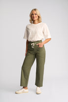 A woman modeling olive green wide-leg cropped pants with a cream drawstring waist, paired with a white textured short-sleeve top and white sneakers, against a light gray studio background.
