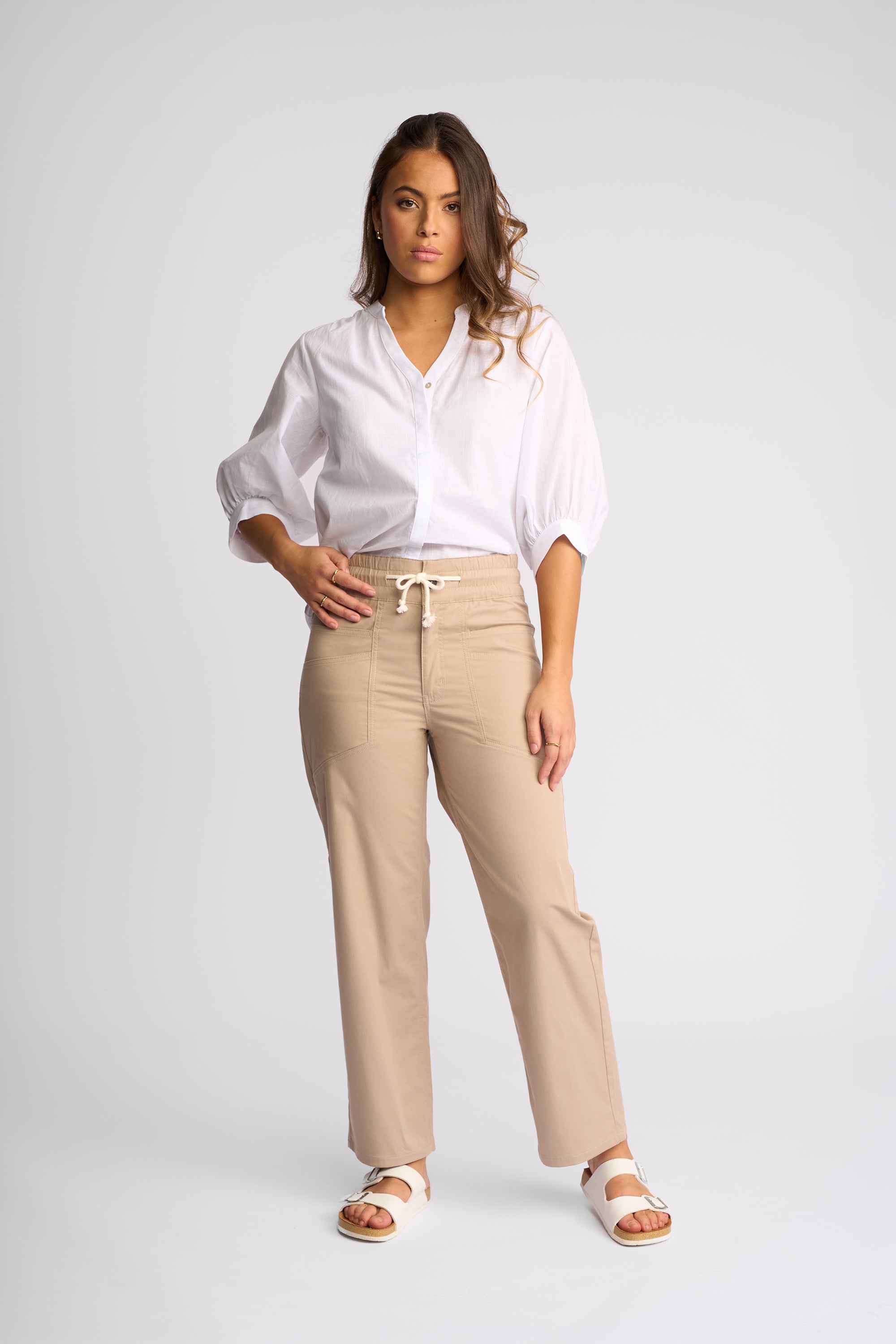 A woman modeling a white short-sleeve blouse and beige wide-leg drawstring pants with white sandals, standing against a light gray studio background.