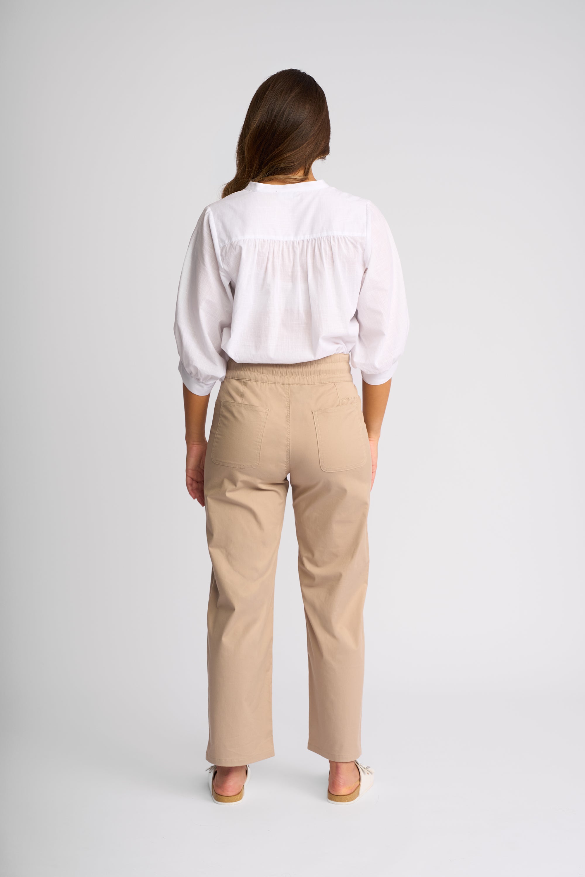 A woman shown from the back wearing a white blouse with a gathered yoke and three-quarter sleeves, paired with straight-leg beige trousers and beige flat sandals, against a light gray studio background.