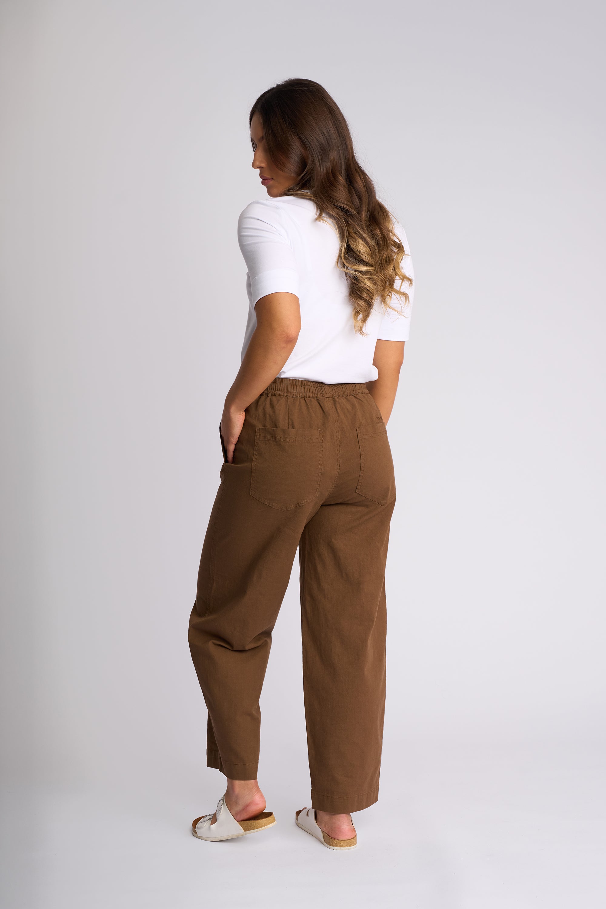 A woman shown from the back wearing brown wide-leg trousers, a white short-sleeve top, and white slide sandals against a light gray studio background.