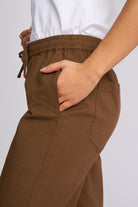 Close-up of a person wearing brown wide-leg pants with an elastic drawstring waist and side pockets, paired with a white top against a light gray background.
