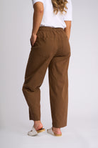 A person shown from the back wearing loose brown cropped pants with a white T-shirt and white slide sandals, photographed against a light gray studio background.