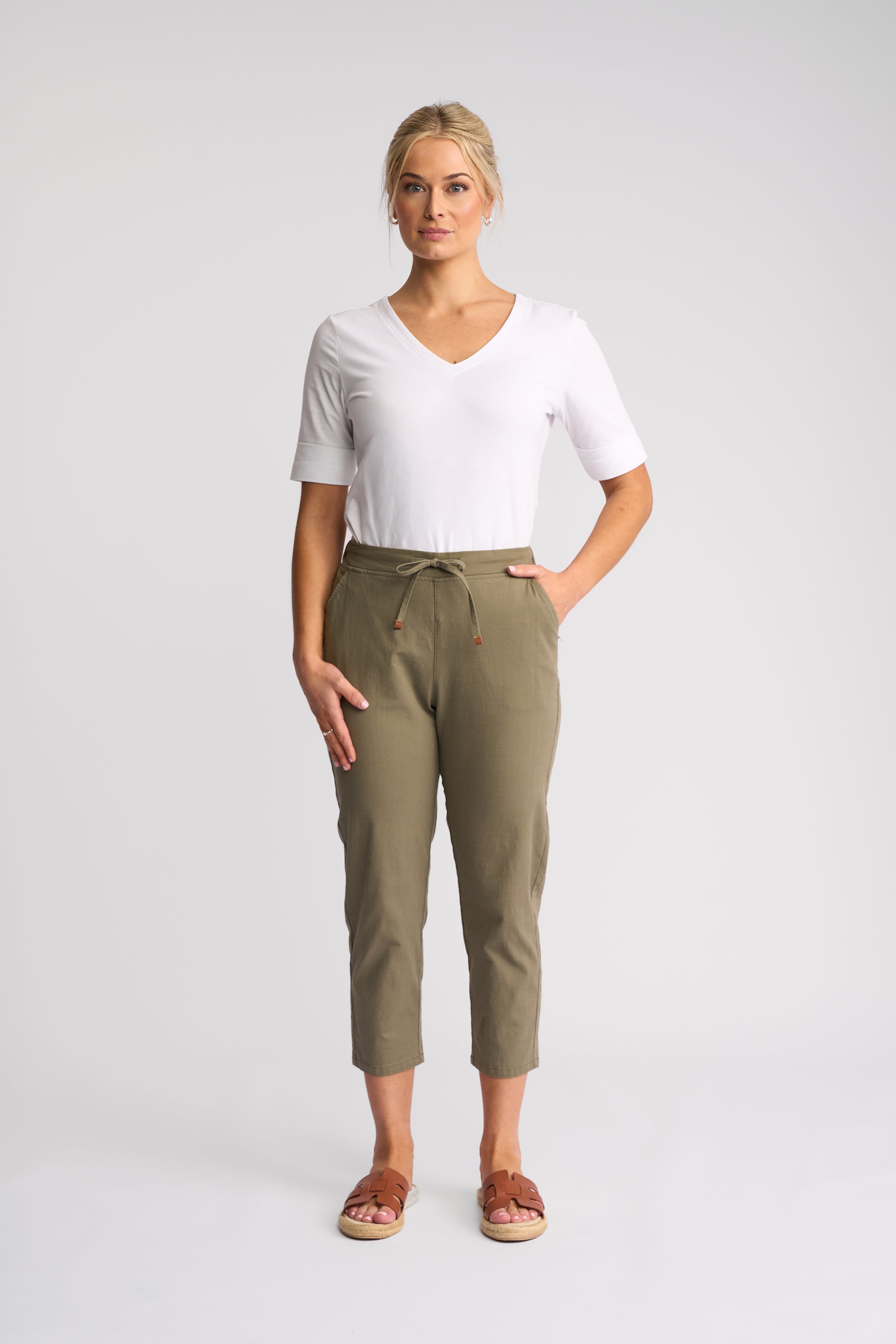 A woman modeling a white short-sleeve V-neck t-shirt and olive green cropped drawstring pants, paired with brown slide sandals, against a light gray studio background.