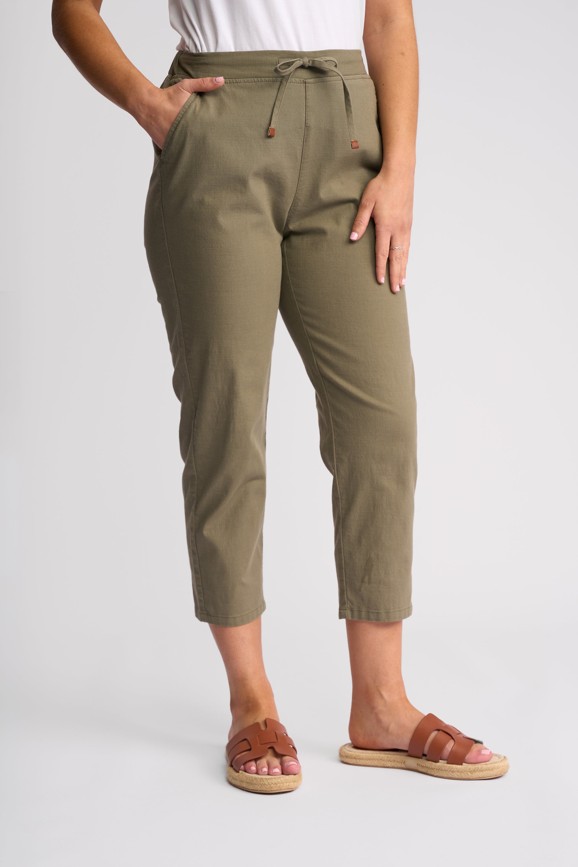 A person wearing olive green cropped drawstring pants with a white top and brown slide sandals, shown from the waist down against a light gray studio background.