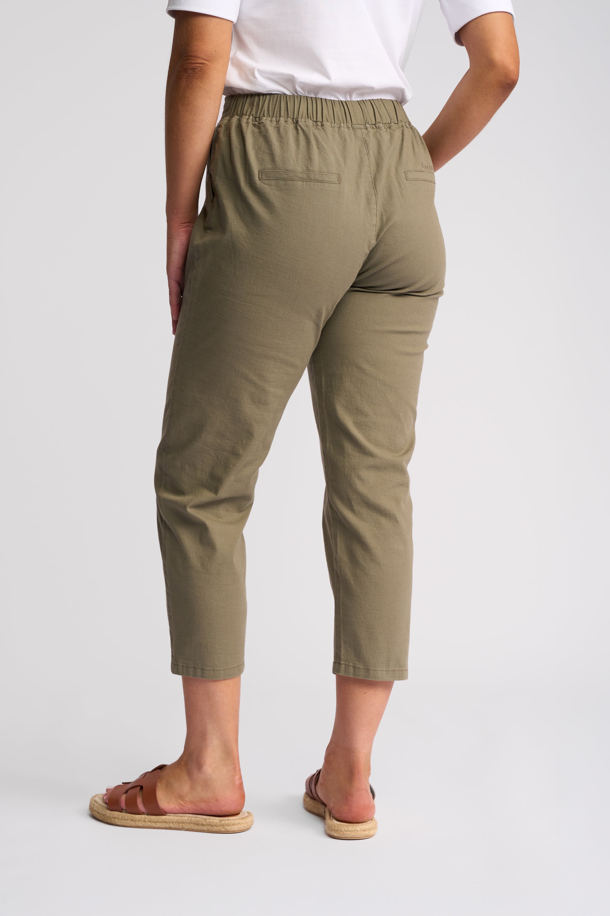 A person shown from the back wearing olive green cropped pants, a white top, and brown slide sandals against a light gray studio background.