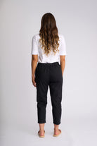 A woman viewed from behind wearing a white short-sleeve top and black cropped pants, standing against a light gray studio background.