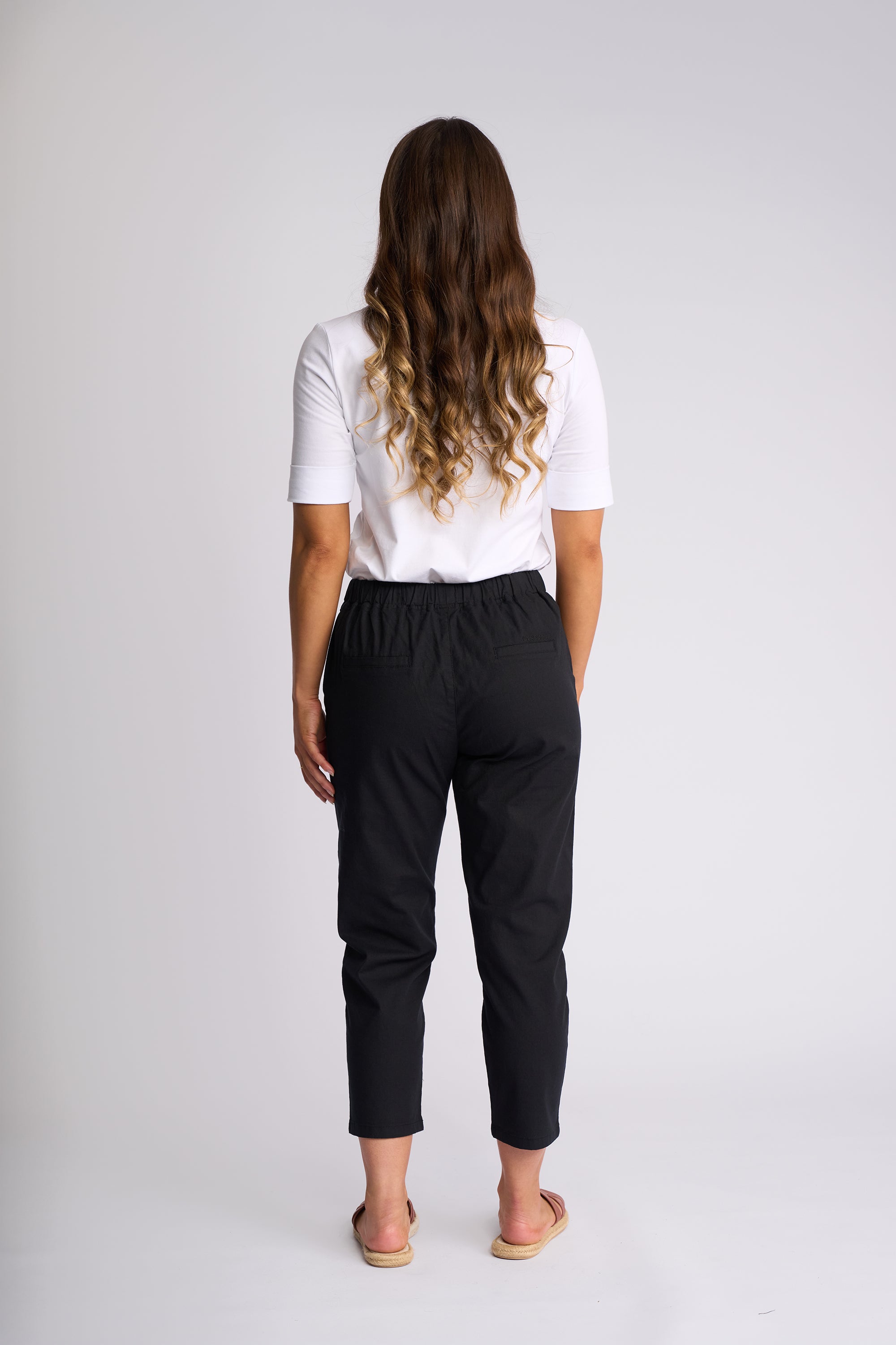 A woman viewed from behind wearing a white short-sleeve top and black cropped pants, standing against a light gray studio background.