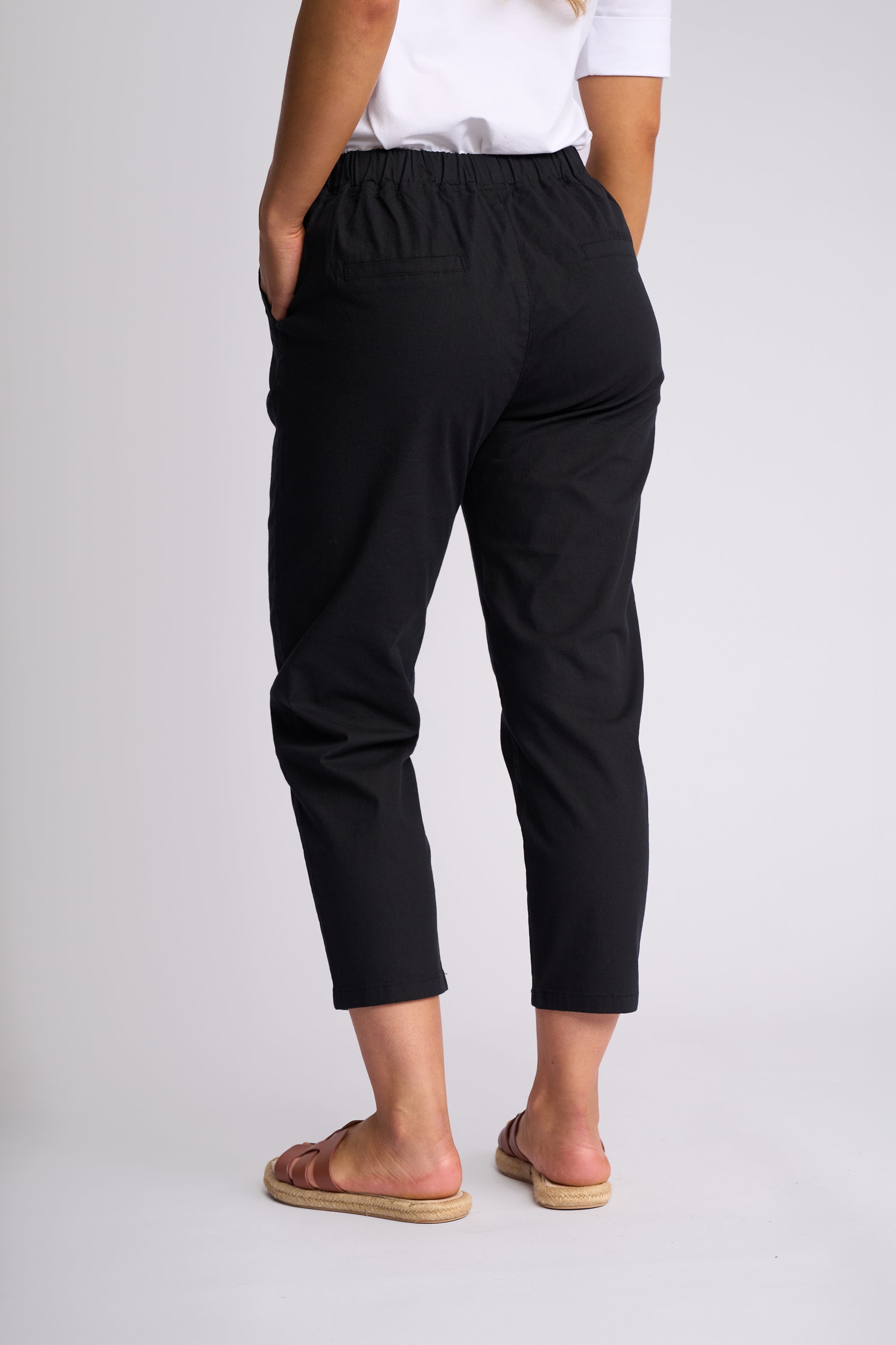 A person shown from the back wearing black cropped pants with an elastic waistband, a white top, and tan slide sandals against a light gray studio background.