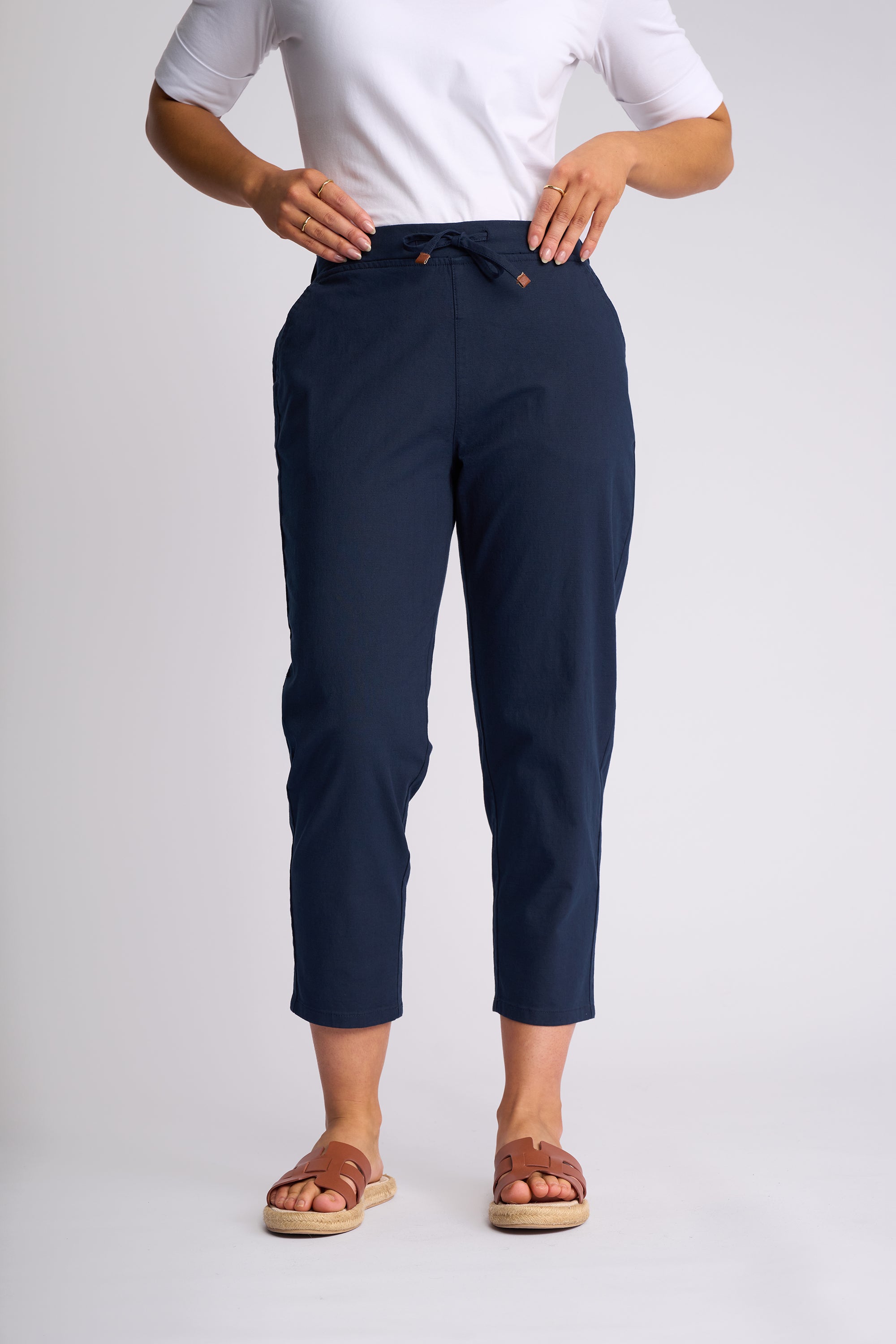 A model wearing navy blue cropped pants with a drawstring waistband, paired with a white t-shirt and brown slide sandals, shown from the waist down on a light gray studio background.