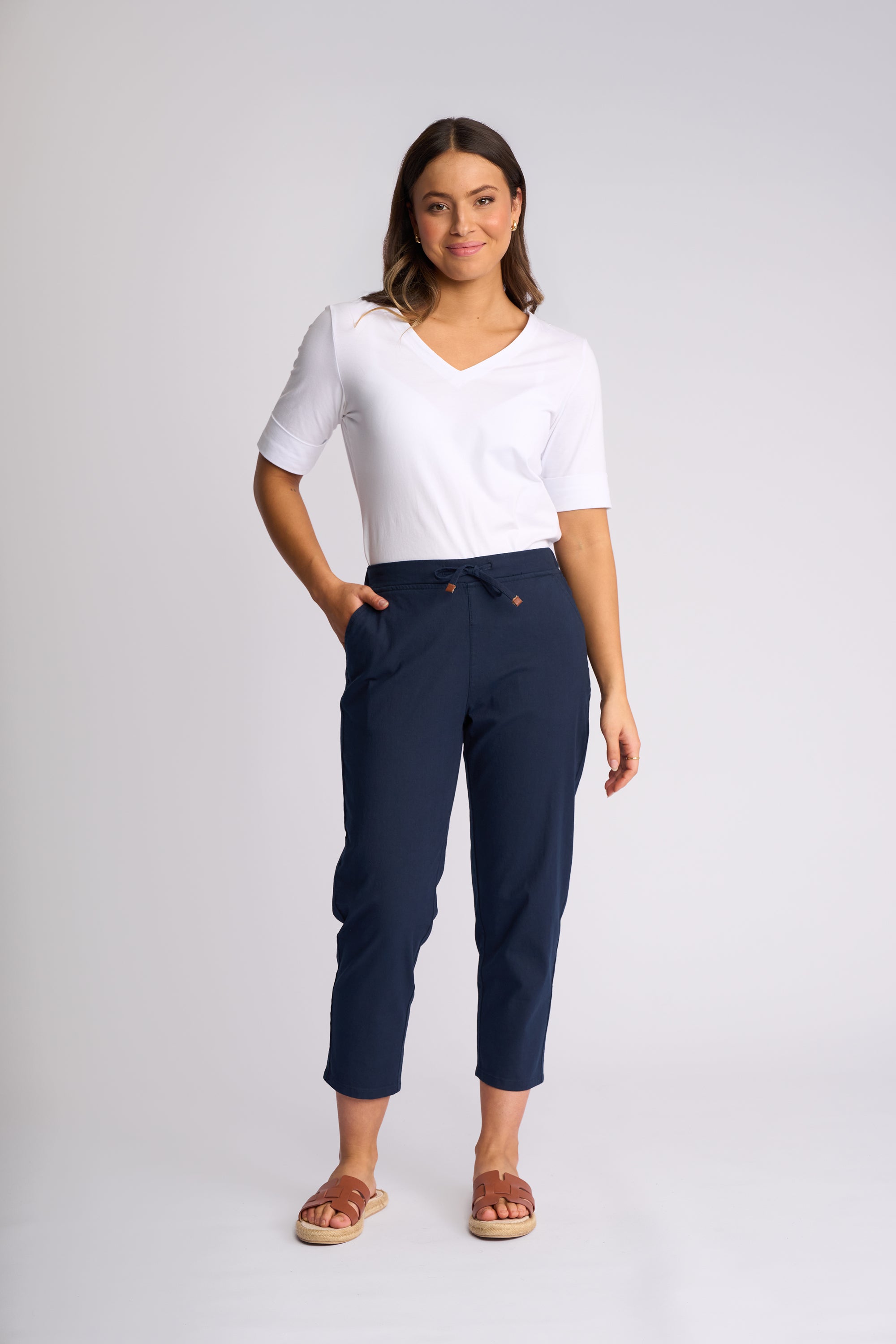 A woman modeling a pair of navy blue cropped pants with a drawstring waist, paired with a white short-sleeve V-neck top and brown slide sandals, against a light gray studio background.
