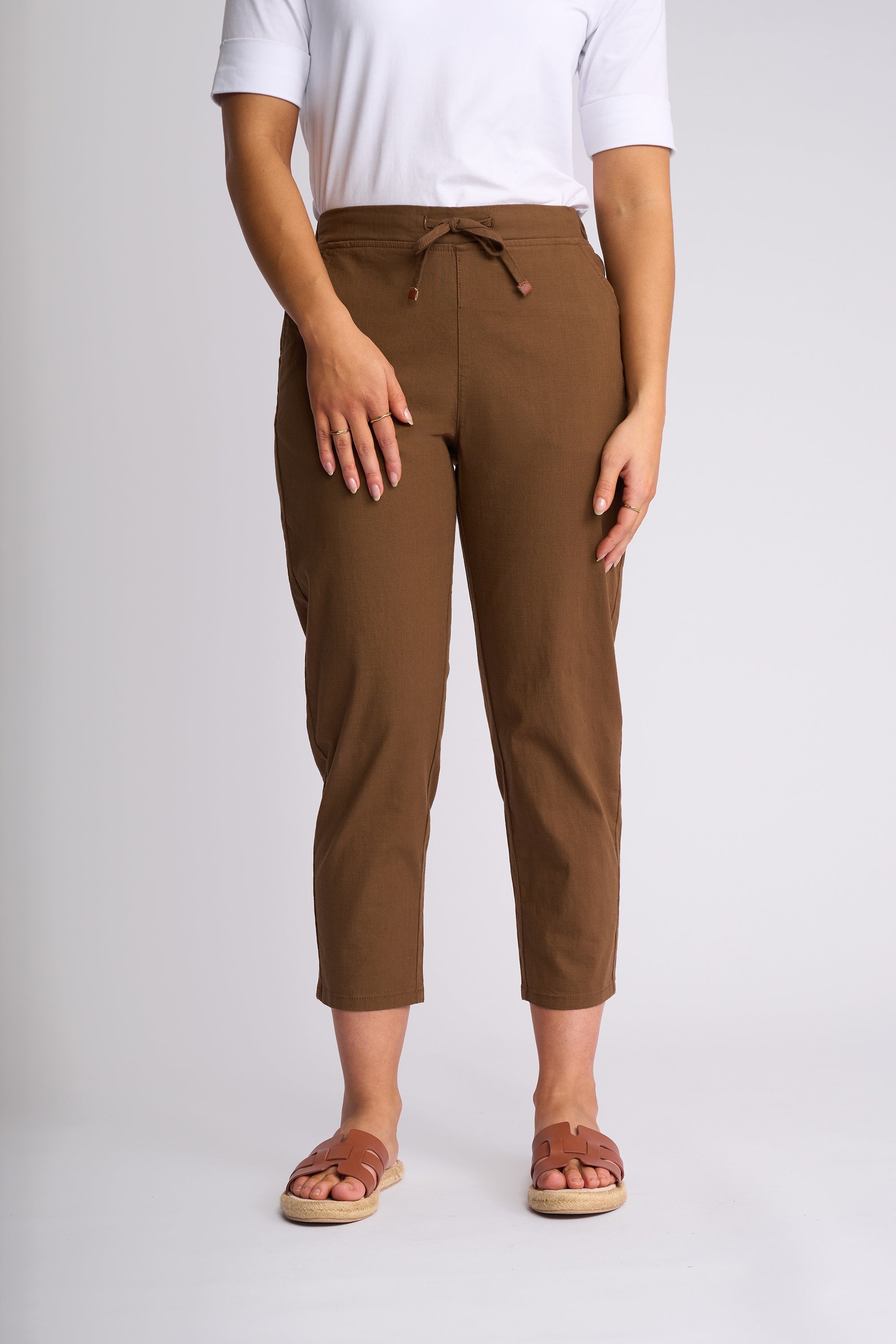 A person wearing brown cropped drawstring pants with a white t-shirt and brown slide sandals, standing against a light gray studio background.
