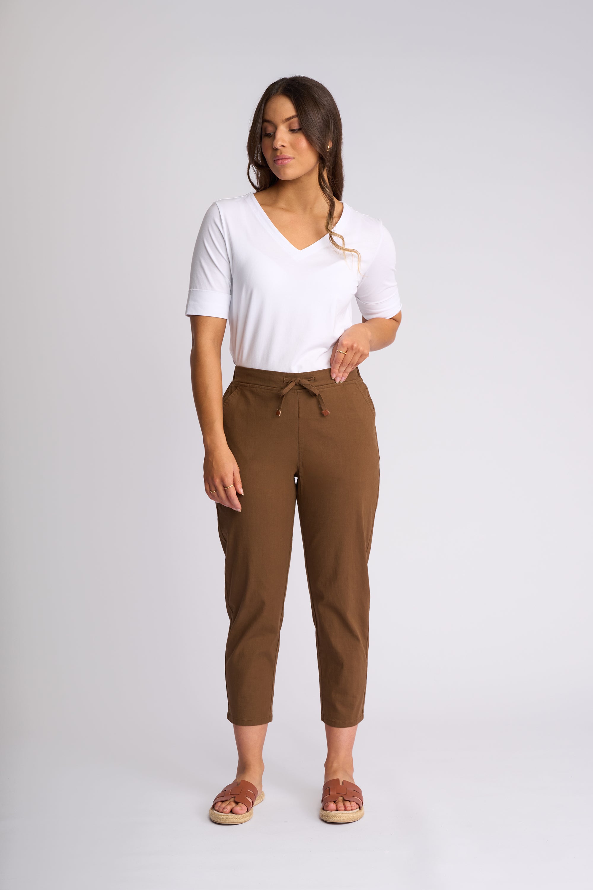 A woman modeling a white short-sleeve V-neck t-shirt and brown cropped drawstring pants against a light gray studio background.