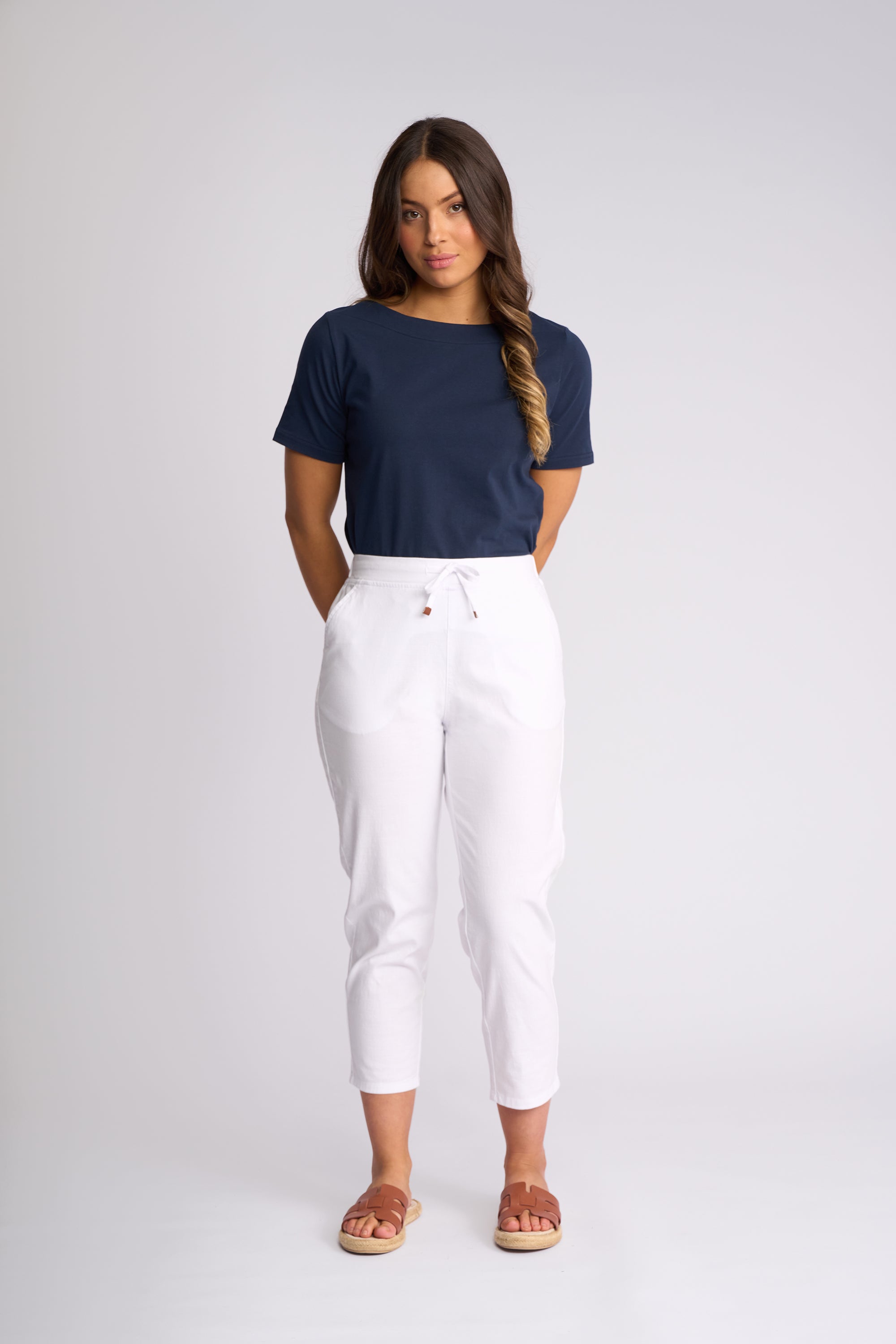 A woman modeling a navy blue short-sleeve crewneck t-shirt and white cropped drawstring pants, paired with brown slide sandals, against a light gray studio background.