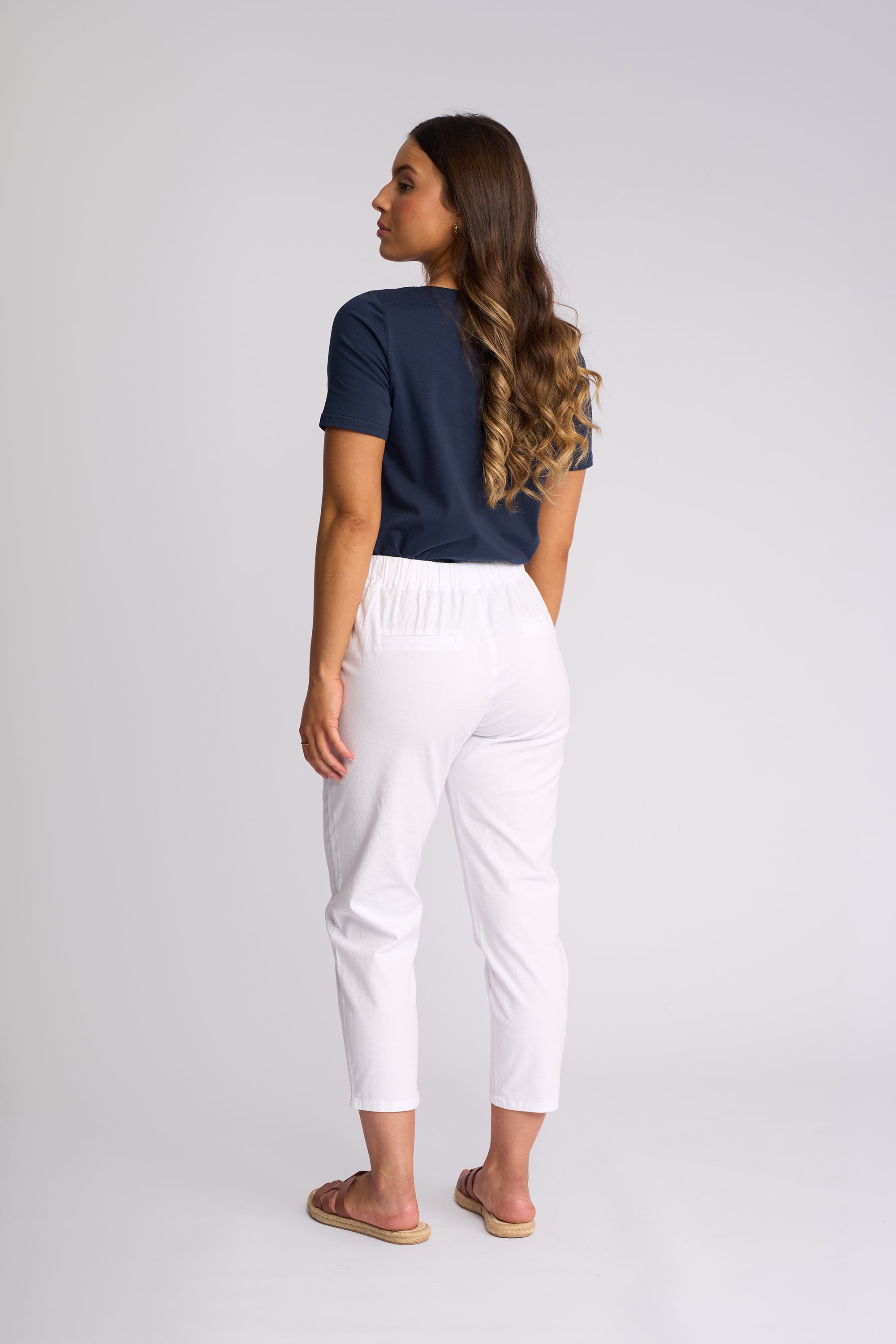 A woman is shown from the back wearing white cropped pants, a navy blue t-shirt, and tan slide sandals against a light gray studio background.