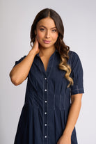 A woman modeling a navy blue short-sleeve button-front shirt dress with white contrast stitching details, set against a plain light gray studio background.