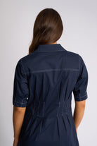 A woman viewed from the back wearing a navy blue short-sleeve shirt dress with white contrast stitching, set against a plain light gray studio background.