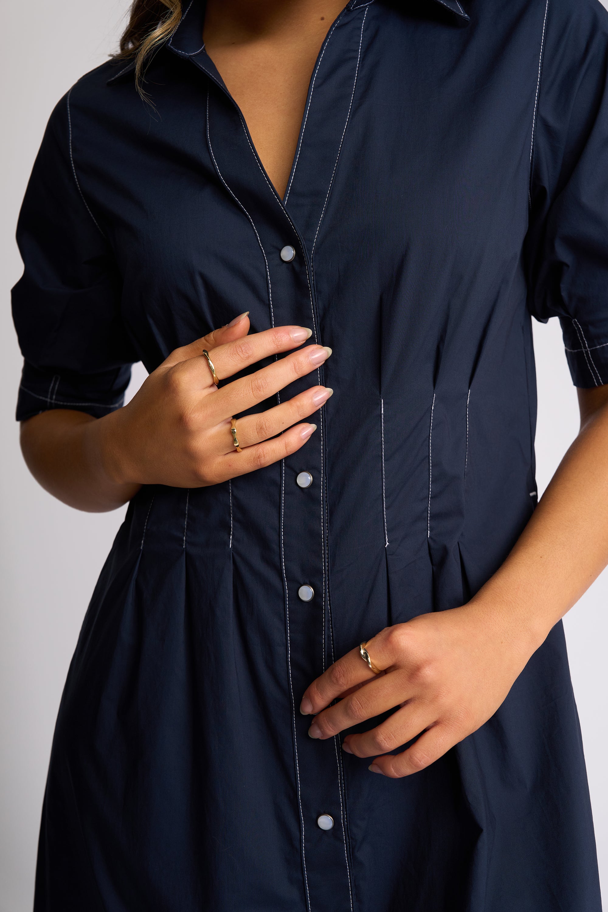 A close-up of a woman wearing a dark navy blue button-up shirt dress with white contrast stitching and white buttons, shown from the shoulders to the hips.